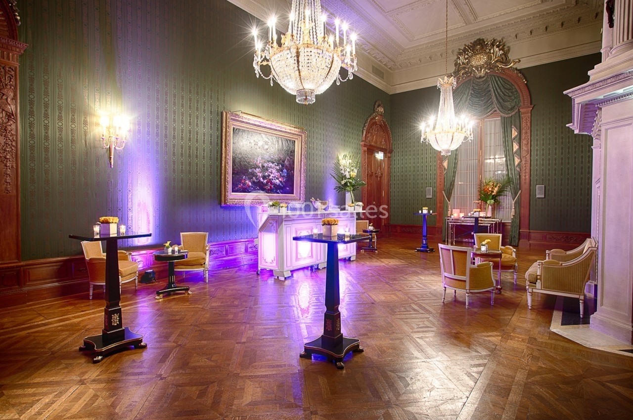 Salle élégante avec parquet, lustres, tables hautes et éclairage tamisé violet, décorée de tableaux et rideaux.
