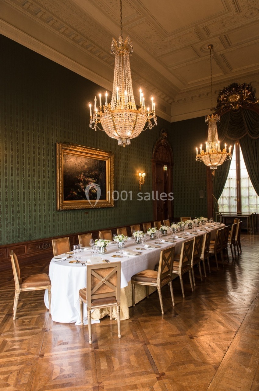 Grande salle élégante avec une longue table dressée, entourée de chaises en bois, éclairée par des lustres en cristal.