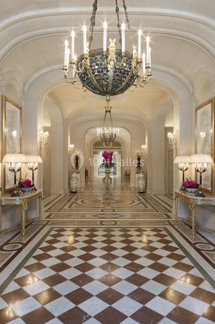 Hall d'entrée luxueux avec sol en damier, lustres imposants, miroirs et tables décorées de fleurs.