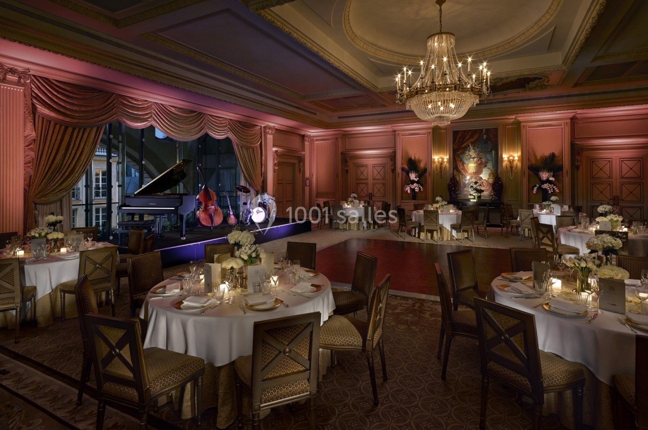 Salle de réception élégante avec tables dressées, chandeliers et scène équipée d'un piano, contrebasse et batterie.