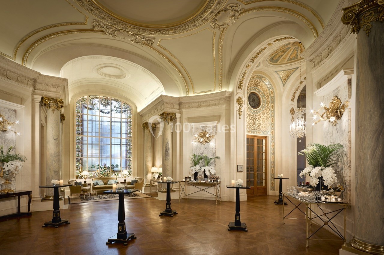Salle élégante avec hauts plafonds ornés, grandes fenêtres, lustres et décorations florales sur des tables.