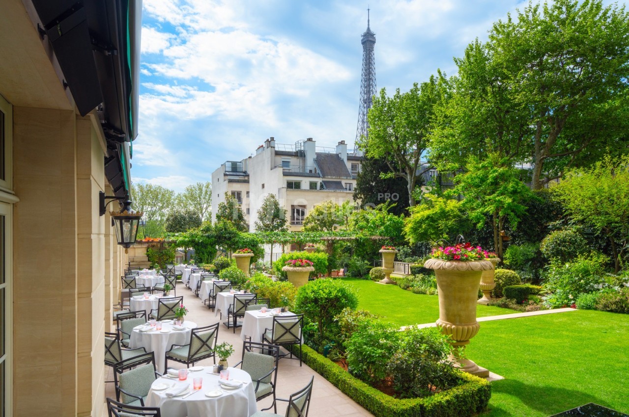 Terrasse d'un restaurant élégant avec des tables dressées, vue sur un jardin verdoyant et la tour Eiffel en arrière-plan.
