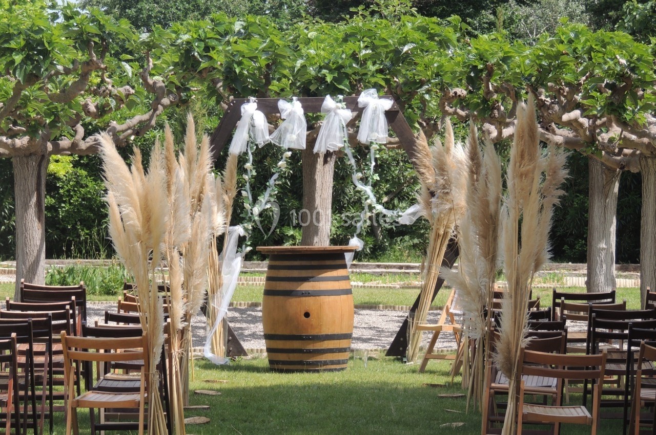 Allée décorée de chaises en bois, d'herbes de la pampa et d'un tonneau sous une arche pour une cérémonie en extérieur.