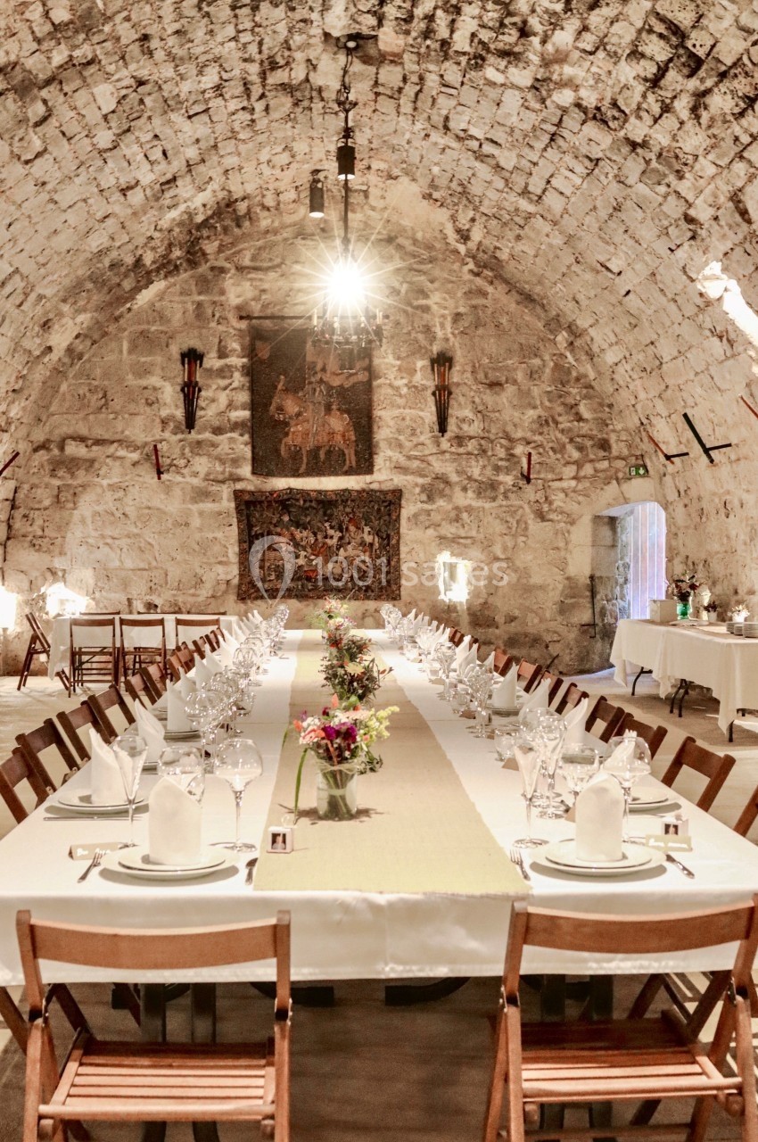 Salle voûtée en pierre avec une longue table dressée pour un repas, décorée de fleurs et éclairée par un lustre.