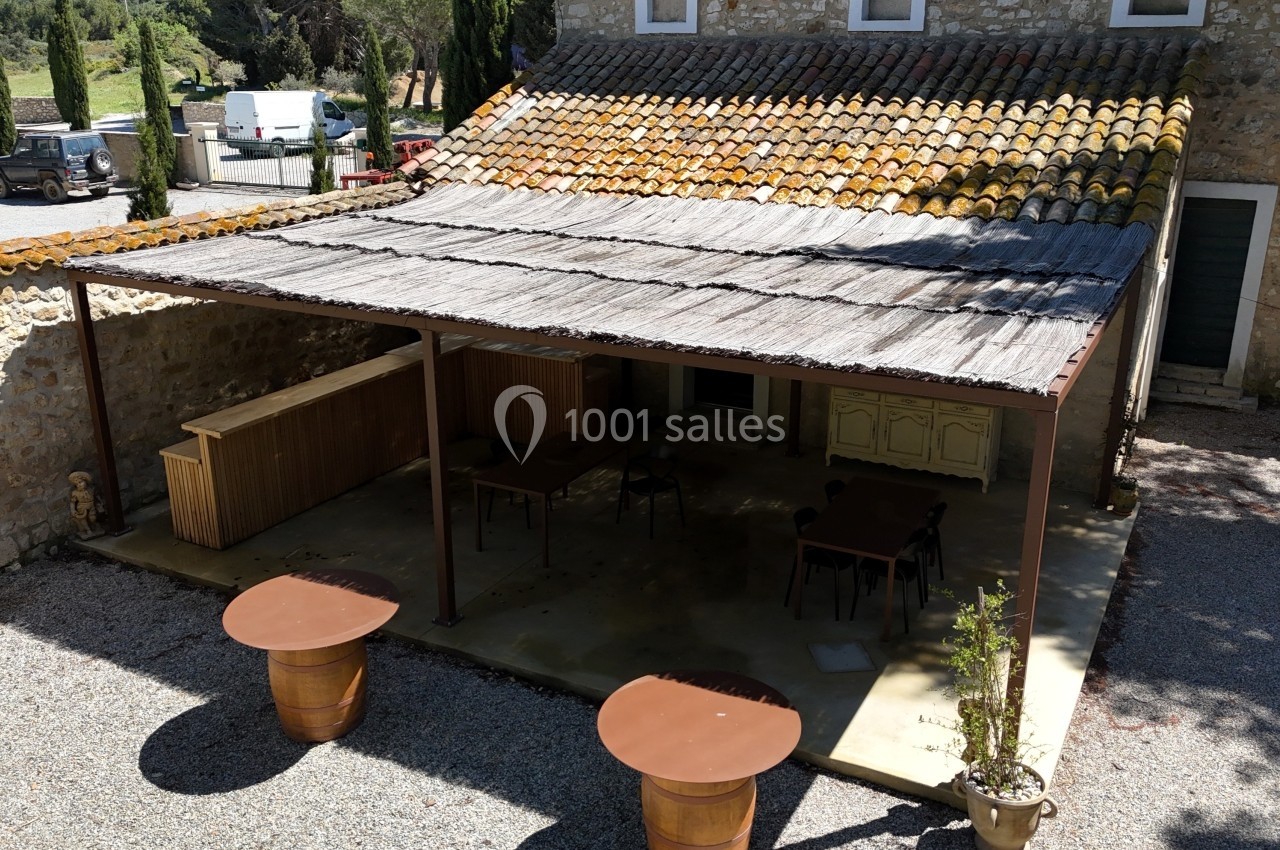 Terrasse couverte avec toit en bois et mobilier extérieur, située devant un bâtiment en pierre dans un cadre rural.