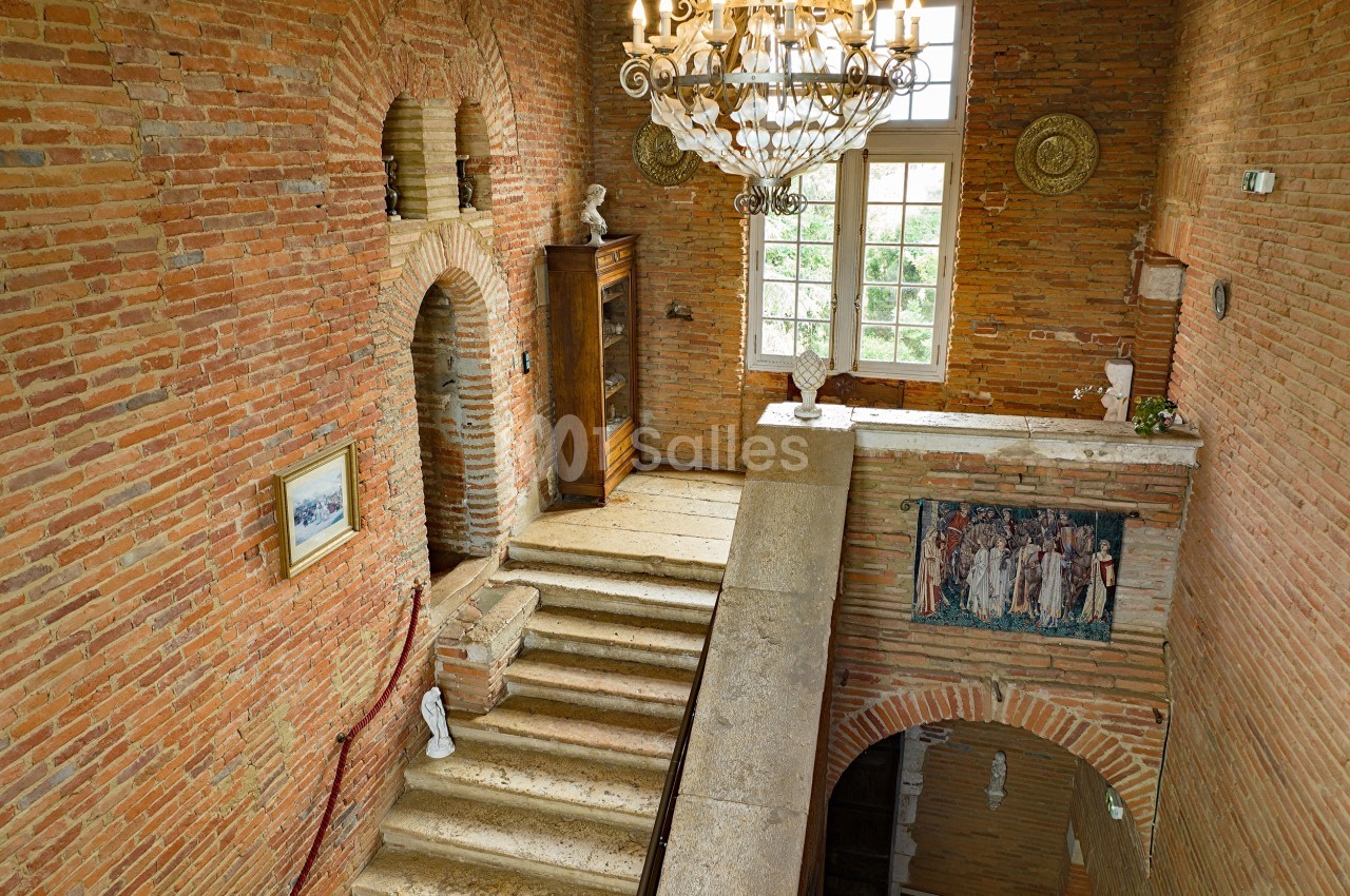 Escalier en pierre dans un intérieur en briques rouges, éclairé par un lustre et une fenêtre avec vue sur la verdure.