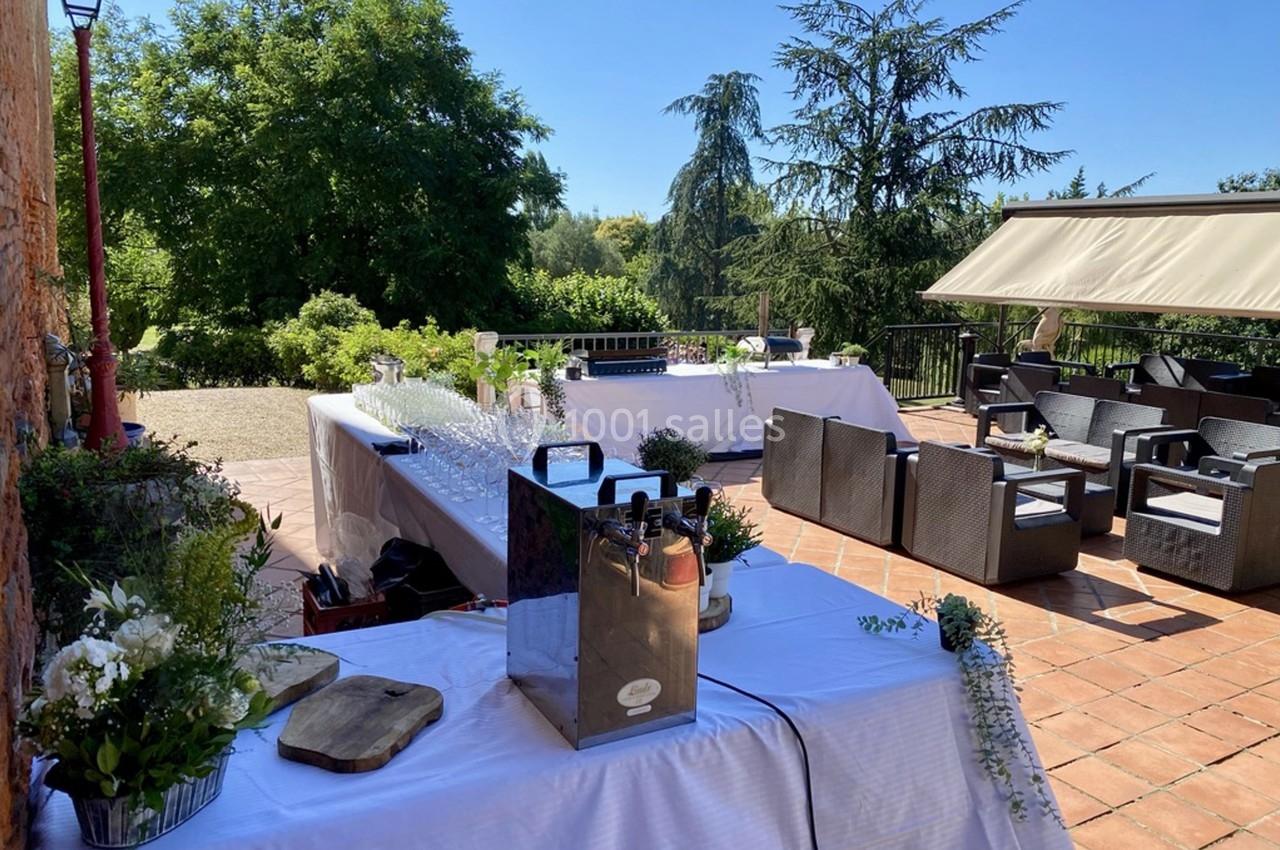 Tables dressées en extérieur avec boissons et décorations florales, entourées de fauteuils et d'arbres sous un ciel dégagé.
