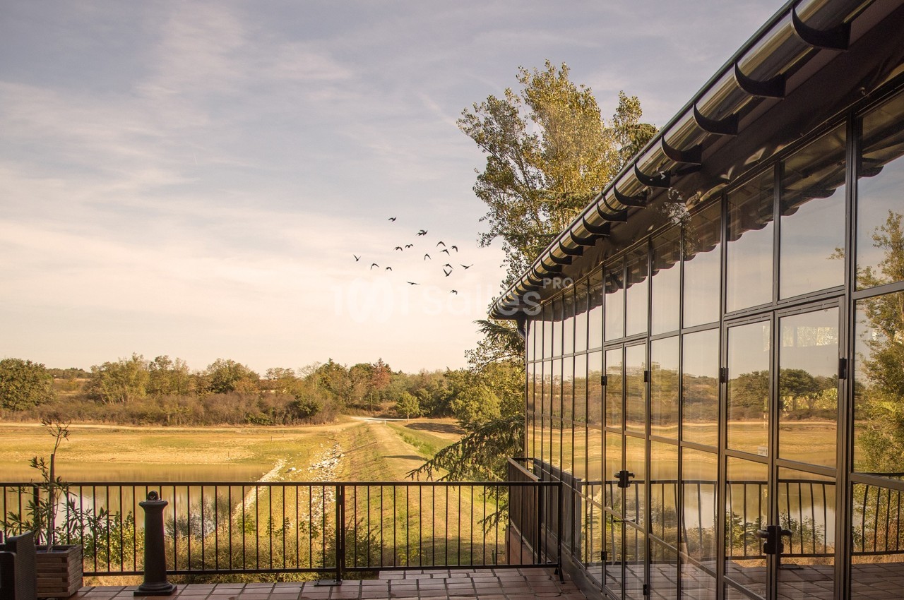 Terrasse en carrelage avec garde-corps, vue sur un paysage naturel avec arbres, champs et un groupe d'oiseaux en vol.