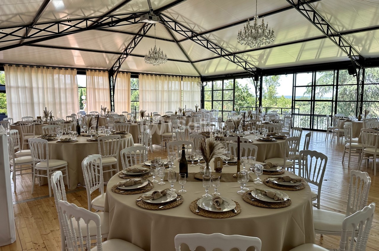 Salle de réception lumineuse avec tables rondes dressées, nappes beiges, chaises blanches et grandes baies vitrées.