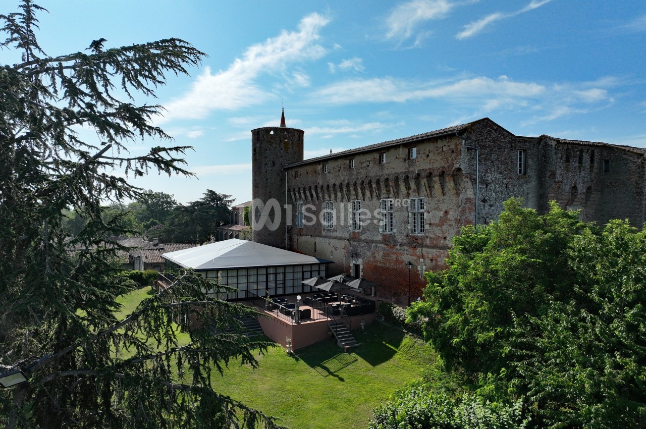 Vue d'un château en pierre avec une tour ronde, une terrasse aménagée et un jardin arboré sous un ciel bleu.