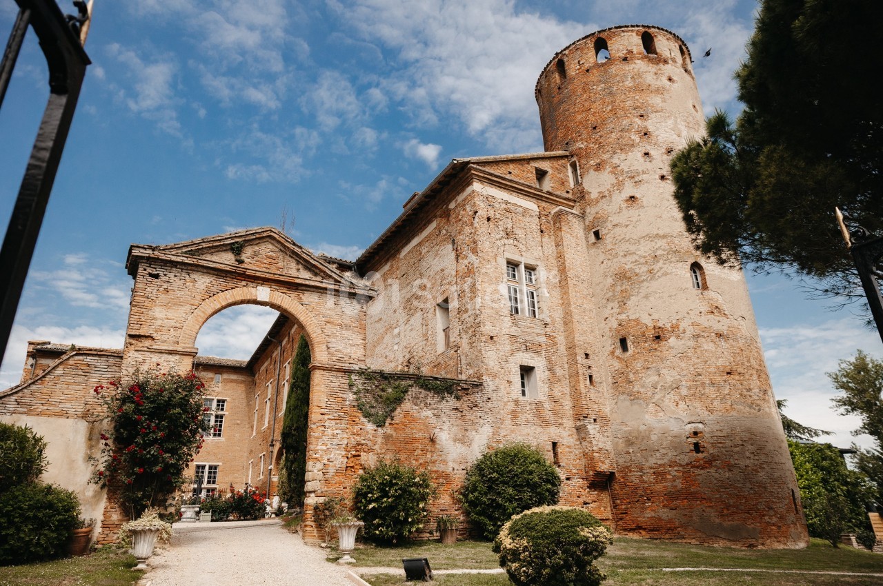 Château en briques avec une tour ronde, une arche et des plantes grimpantes sous un ciel partiellement nuageux.