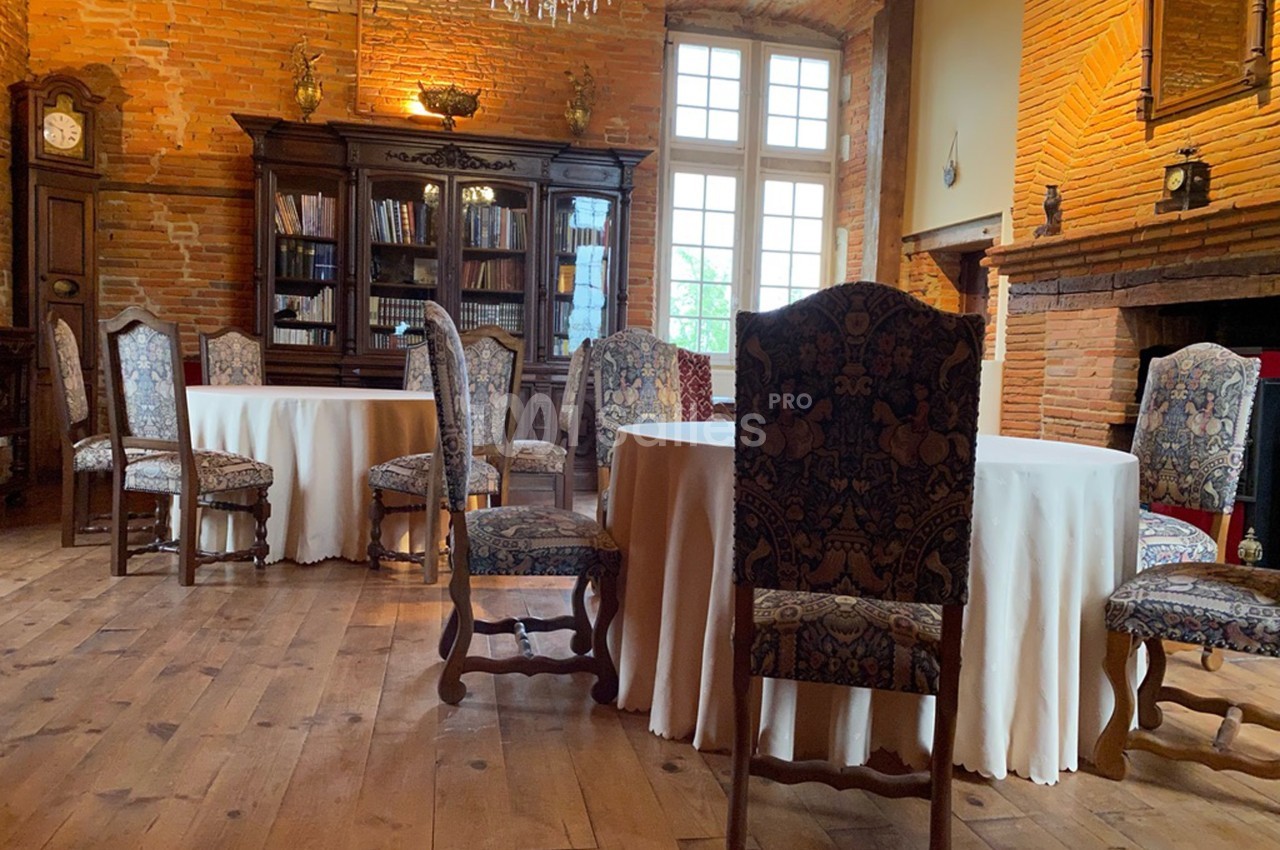 Salle avec des tables rondes couvertes de nappes blanches, chaises en bois sculpté et murs en briques apparentes.