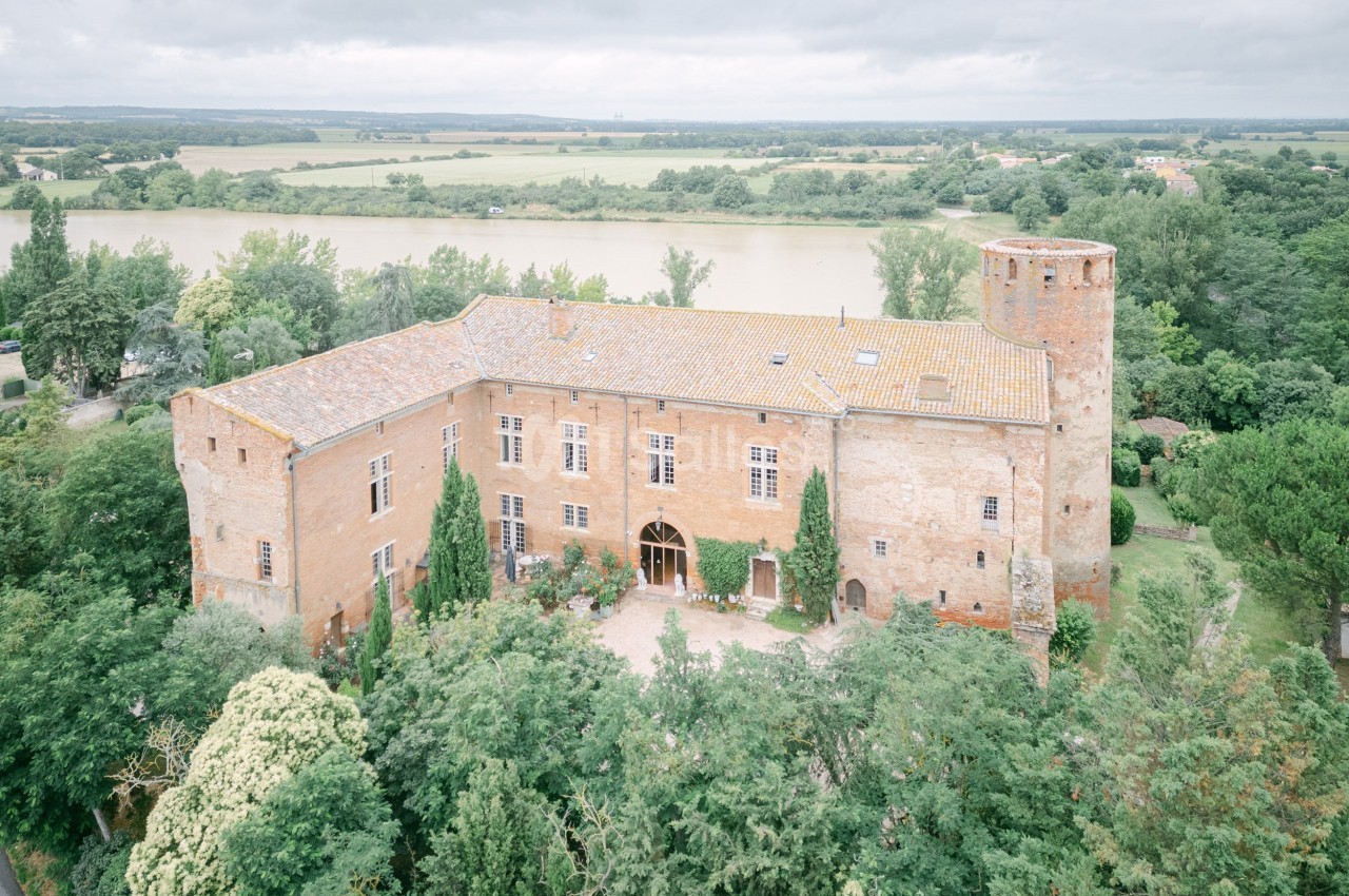 Vue aérienne d'un château en briques rouges entouré de végétation, avec une rivière et des champs en arrière-plan.