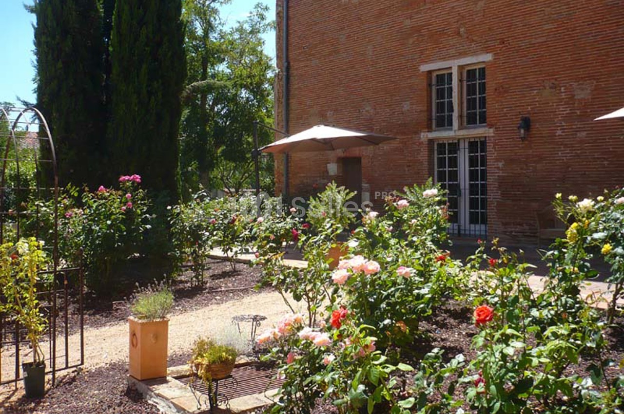 Jardin fleuri avec rosiers, allées en gravier, parasols et façade en briques rouges en arrière-plan.
