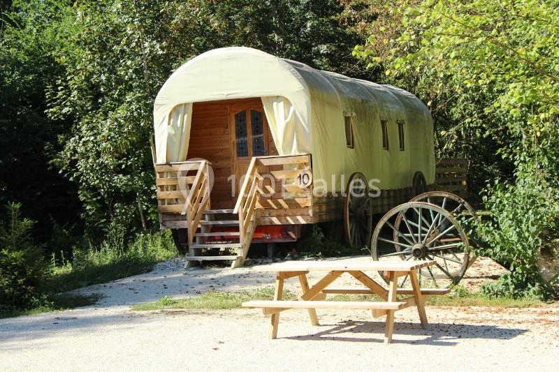 Roulotte en bois avec toit arrondi, entourée de végétation, avec une table de pique-nique et une roue ancienne au premier…