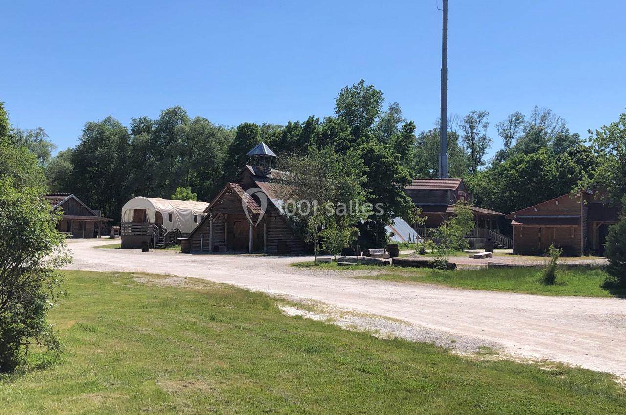 Village en plein air avec bâtiments en bois, une roulotte et une antenne, entouré de verdure sous un ciel dégagé.