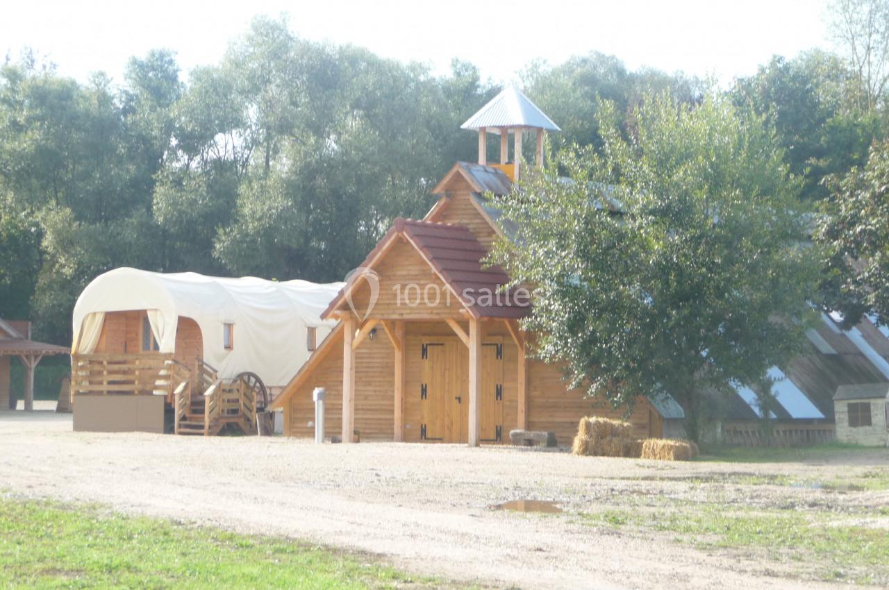 Bâtiments en bois de style western avec une roulotte et un arbre, entourés de verdure.