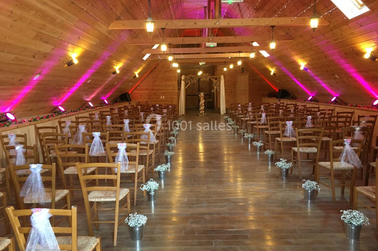 Salle en bois décorée pour une cérémonie, avec des rangées de chaises ornées de rubans blancs et des fleurs au sol.