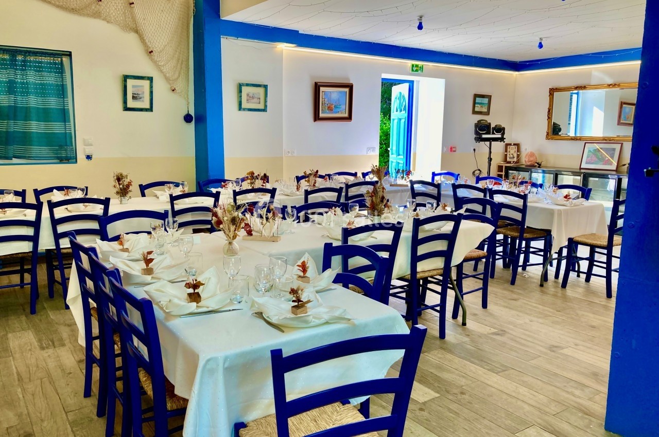 Salle de restaurant décorée en bleu et blanc avec des tables dressées pour un repas, ambiance lumineuse et maritime.