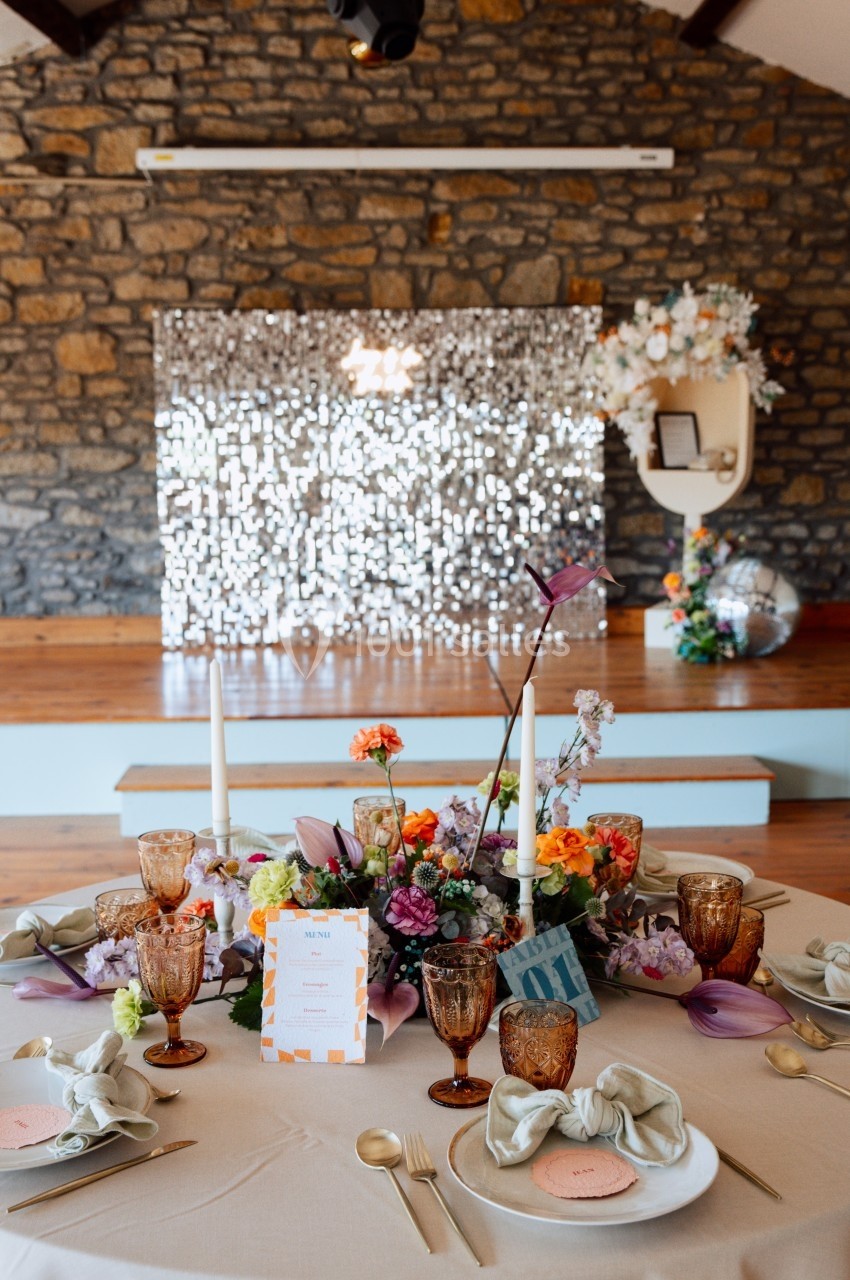 Table décorée pour un événement avec fleurs colorées, bougies, vaisselle élégante et fond en pierres et paillettes.