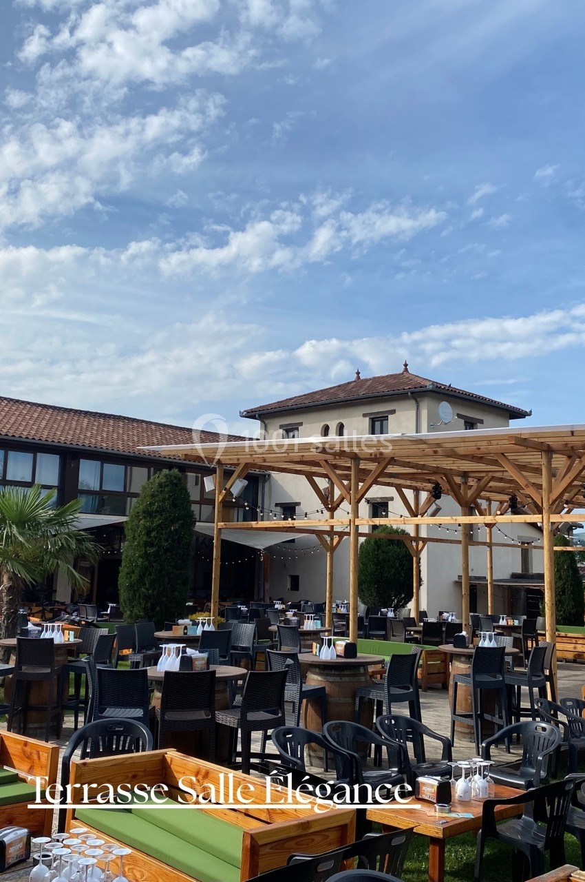 Terrasse extérieure avec tables dressées, pergola en bois et bâtiment à toit en tuiles en arrière-plan.
