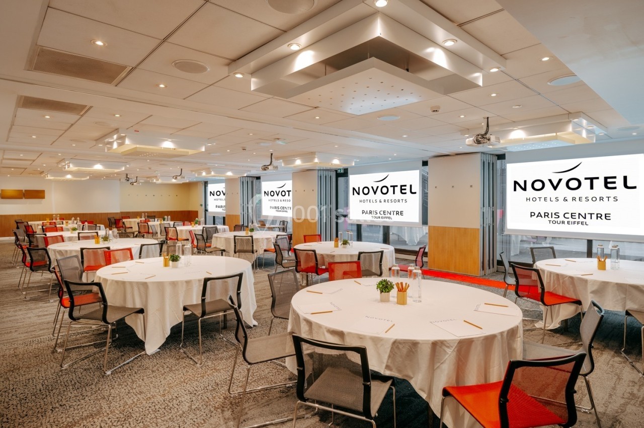 Salle de conférence lumineuse avec tables rondes dressées, chaises colorées et écrans affichant le logo Novotel.