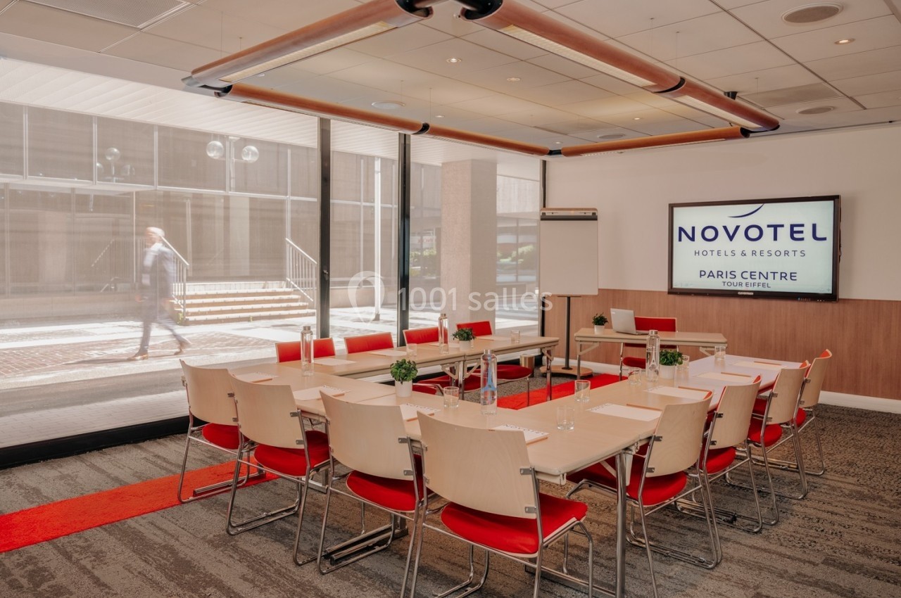 Salle de réunion lumineuse avec tables disposées en U, chaises rouges et écran affichant ’Novotel Paris Centre’.
