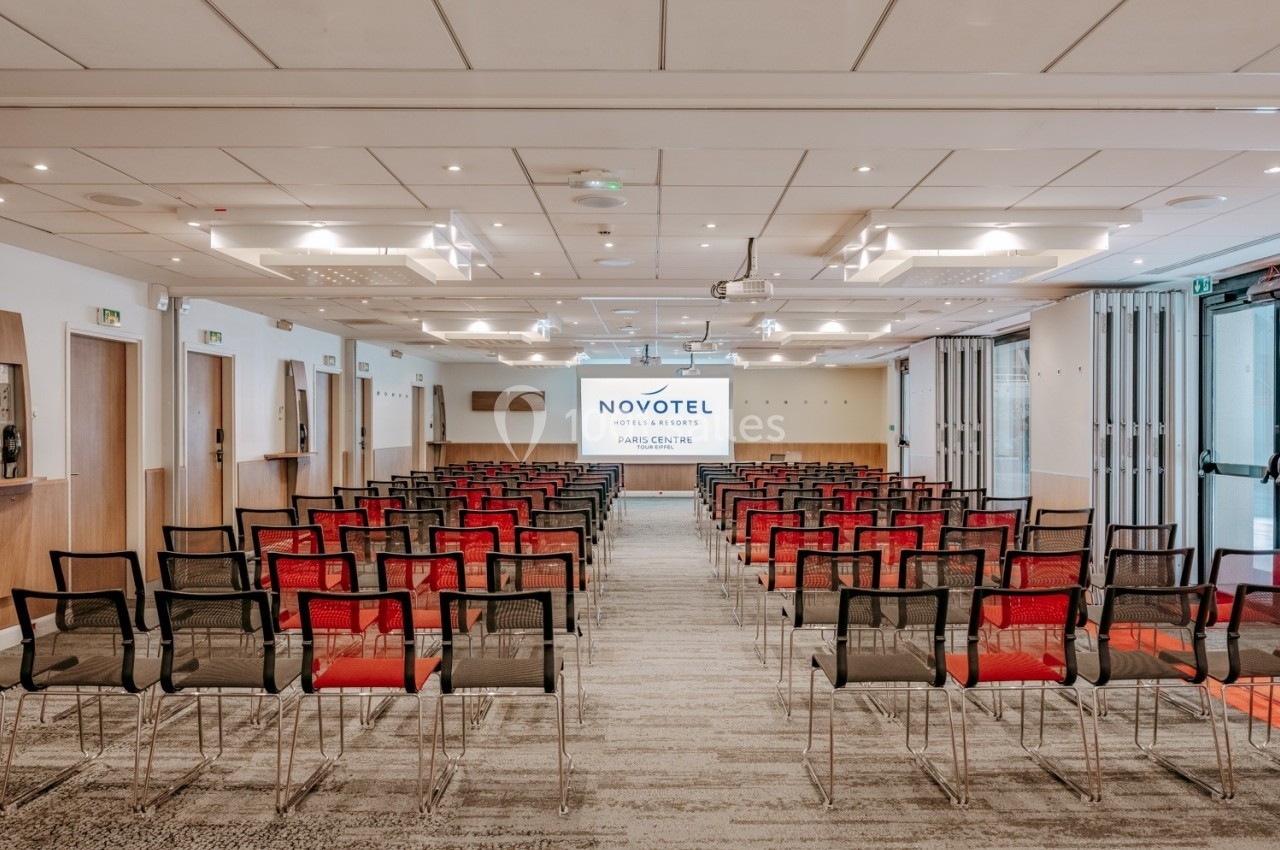 Salle de conférence lumineuse avec des rangées de chaises noires et rouges, écran de projection au fond.