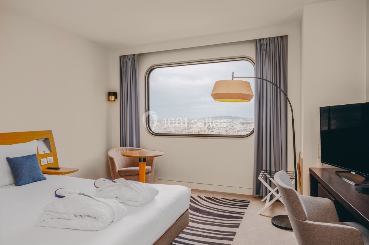 Chambre d'hôtel lumineuse avec lit double, fauteuils, bureau, télévision et grande fenêtre offrant une vue sur la ville.