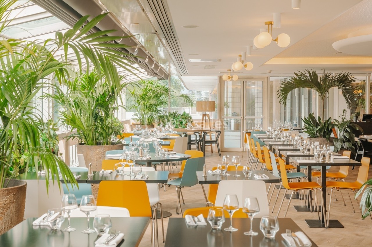 Salle de restaurant lumineuse avec des tables dressées, des chaises colorées et une décoration végétale abondante.