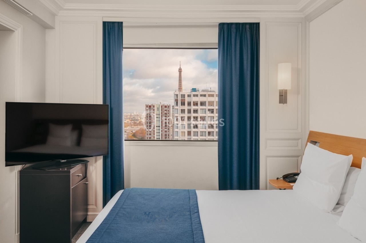Chambre d'hôtel avec lit double, télévision, et vue sur la Tour Eiffel à travers une fenêtre encadrée de rideaux bleus.