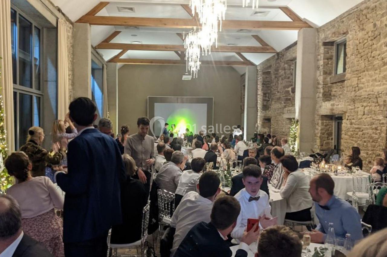 Salle de réception avec des invités assis à des tables, éclairage tamisé et scène illuminée en arrière-plan.