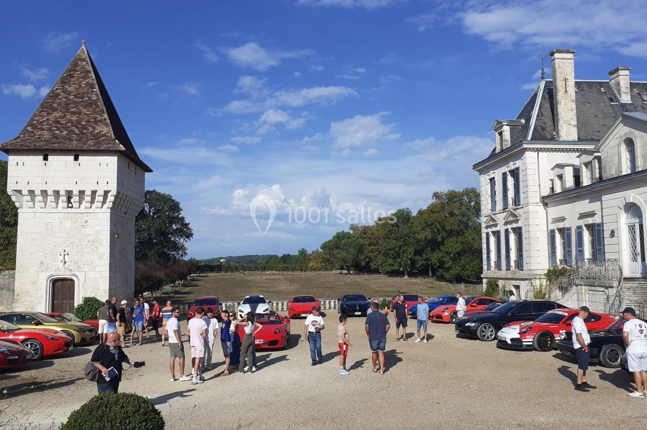 Rassemblement de voitures de sport devant un château et une tour, avec des visiteurs en plein air.