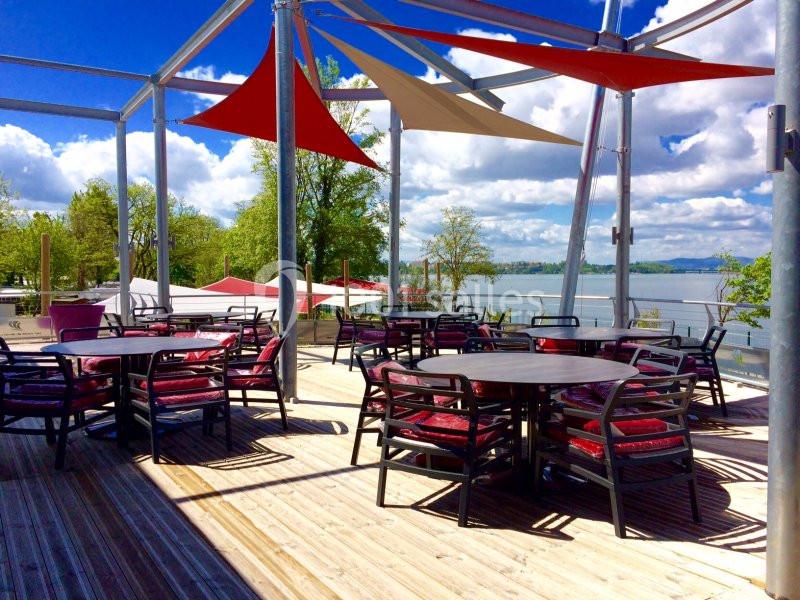 Terrasse en bois avec tables et chaises, surplombant un lac sous un ciel partiellement nuageux.