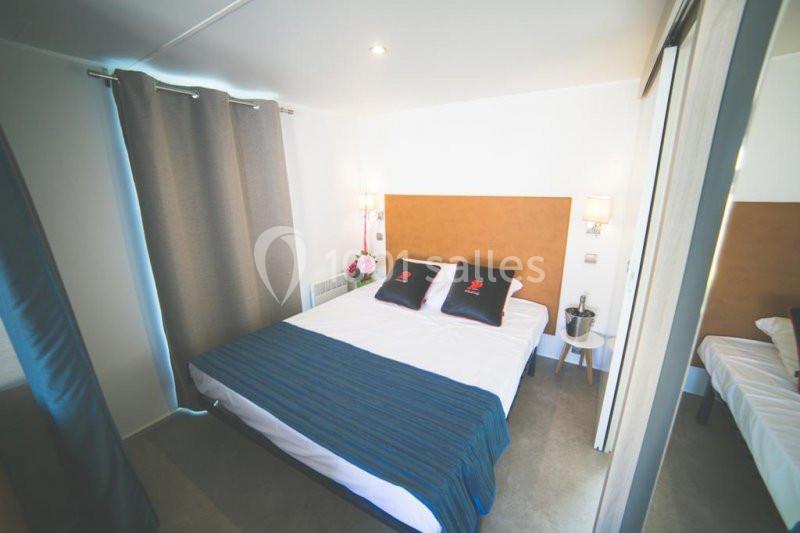 Chambre lumineuse avec un lit double, draps bleus, coussins noirs, rideaux gris et une décoration simple.