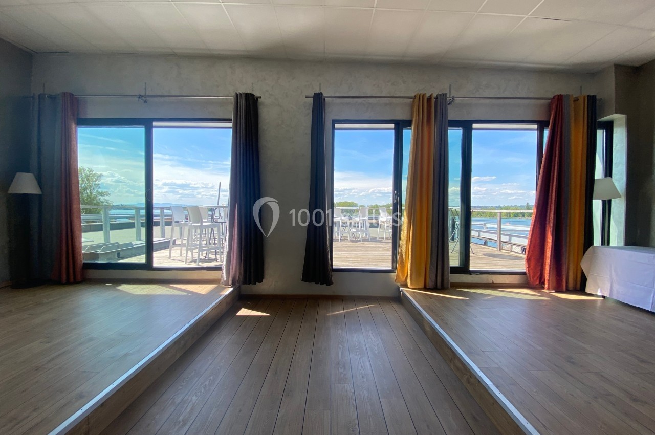 Deux grandes baies vitrées avec rideaux donnant sur une terrasse ensoleillée et une vue sur un plan d'eau.