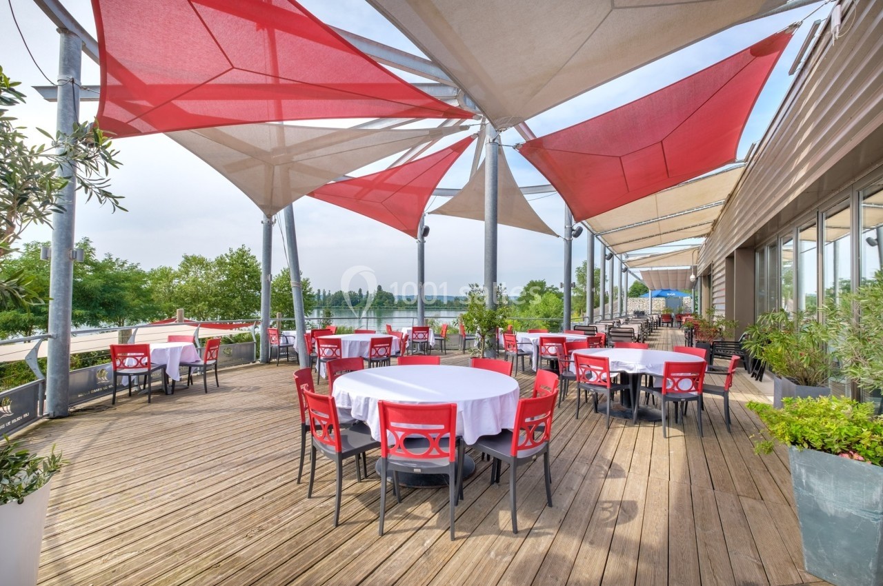 Terrasse en bois avec tables rondes dressées, chaises rouges et grandes toiles d'ombrage, vue sur un paysage verdoyant.