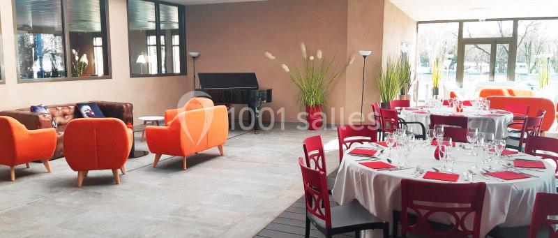 Salle lumineuse avec tables dressées pour un repas, fauteuils orange, piano noir et plantes décoratives.