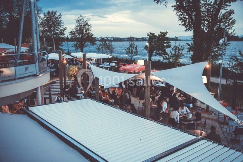 Terrasse extérieure animée en bord de lac, avec des tables, des parasols et des convives sous un ciel nuageux.