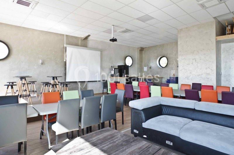 Salle lumineuse avec tables, chaises colorées, canapé, écran de projection et murs en béton clair.