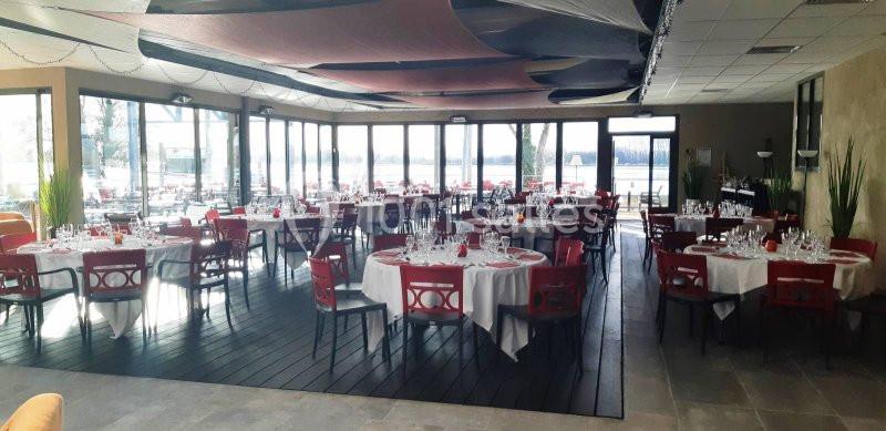 Salle de réception lumineuse avec grandes baies vitrées, tables rondes dressées et chaises rouges.
