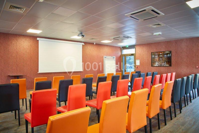 Salle de conférence avec des chaises colorées alignées face à un écran de projection et un tableau blanc.