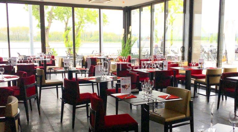 Salle de restaurant lumineuse avec grandes baies vitrées, tables dressées et vue sur un paysage arboré.