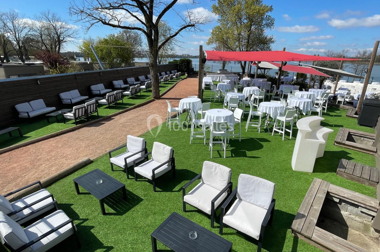 Terrasse extérieure aménagée avec tables rondes, chaises blanches, fauteuils et parasols, entourée de verdure et d'arbres.
