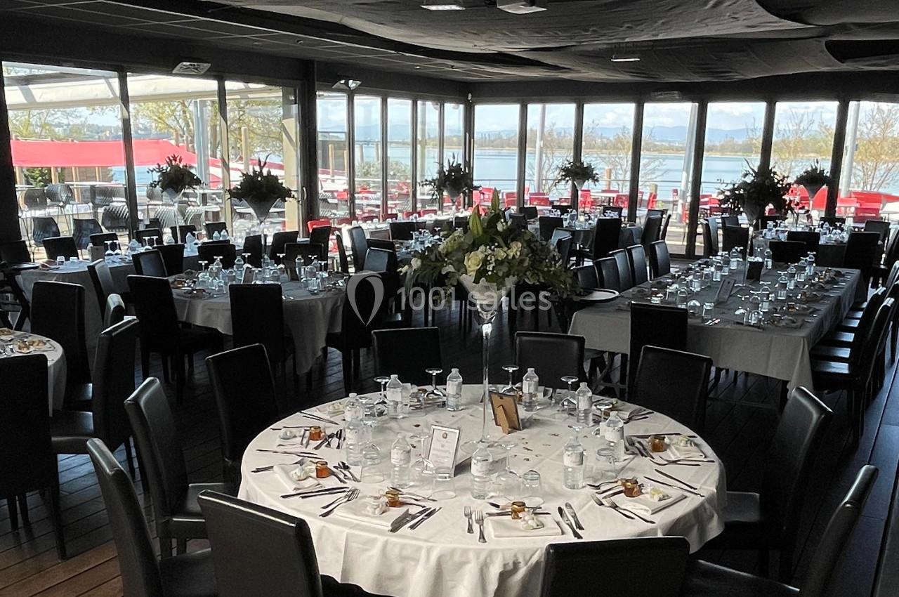 Salle de réception élégamment dressée avec des tables rondes, nappes blanches et vue sur un espace extérieur ensoleillé.