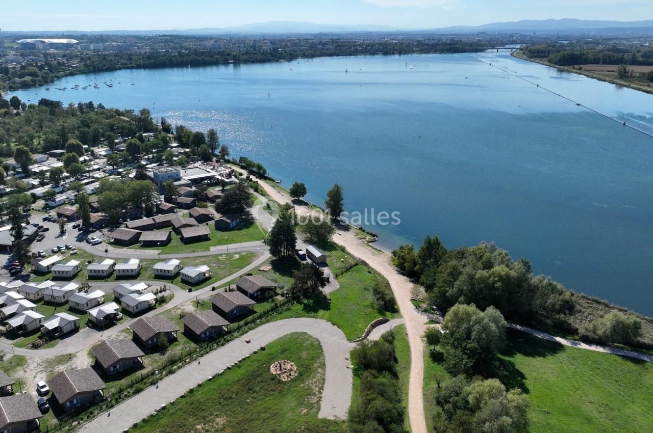Vue aérienne d'un lac entouré de verdure, avec des habitations et un chemin bordant la rive.