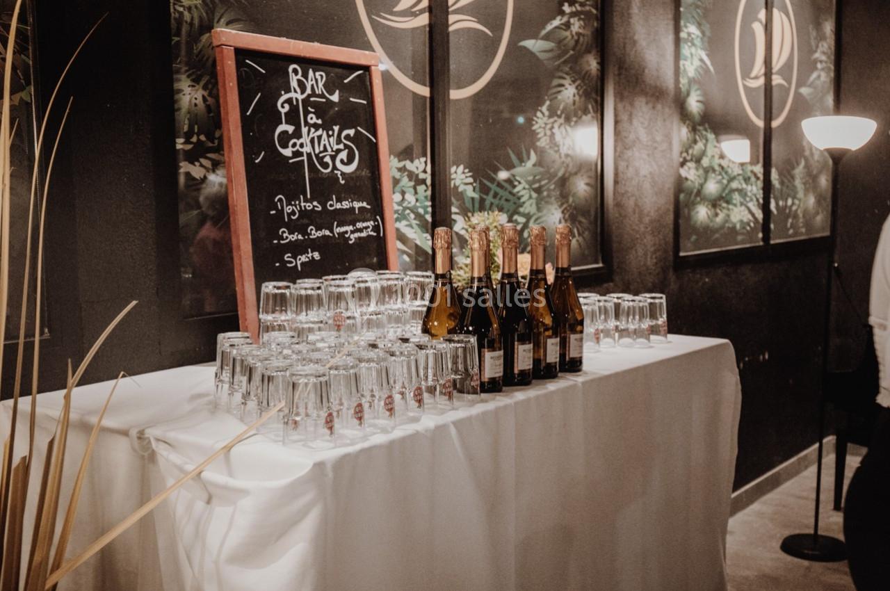 Table dressée avec des bouteilles de champagne, des verres et un tableau indiquant des cocktails dans un décor élégant.