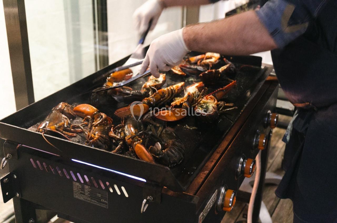 Personne en train de griller des homards sur une plancha, avec des pinces et des gants pour manipuler les crustacés.