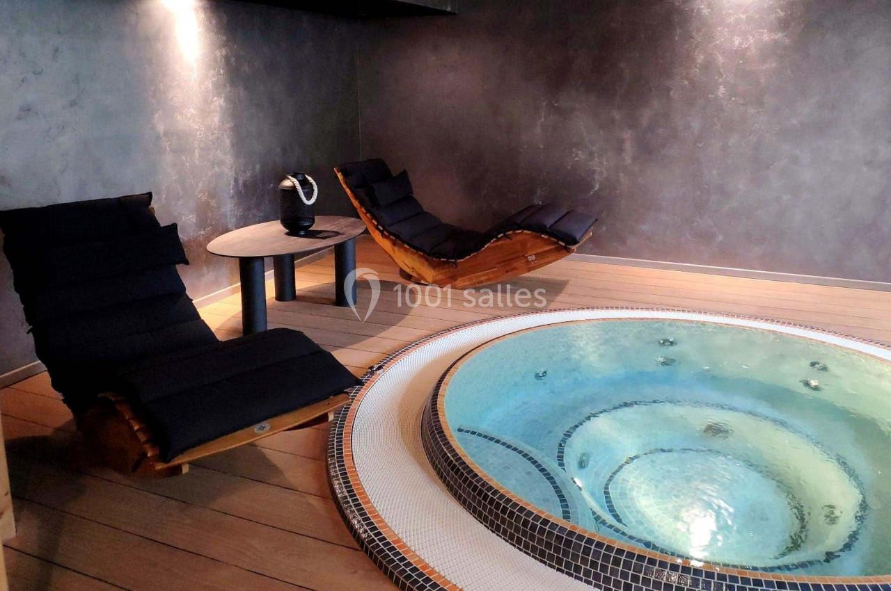 Bain à remous entouré de deux chaises longues en bois dans une pièce au mur sombre et éclairage tamisé.