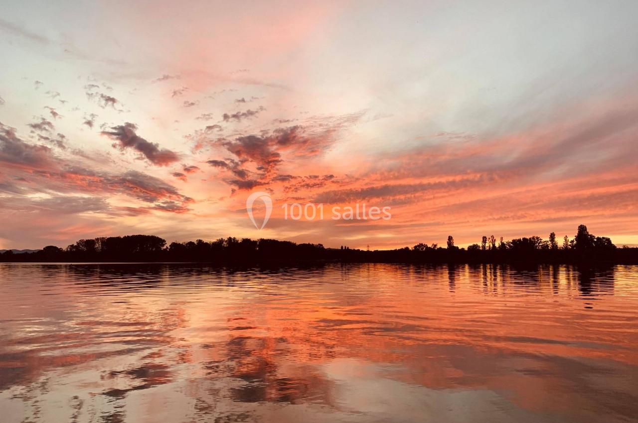 Coucher de soleil aux teintes orangées et roses se reflétant sur un lac calme bordé d'arbres.